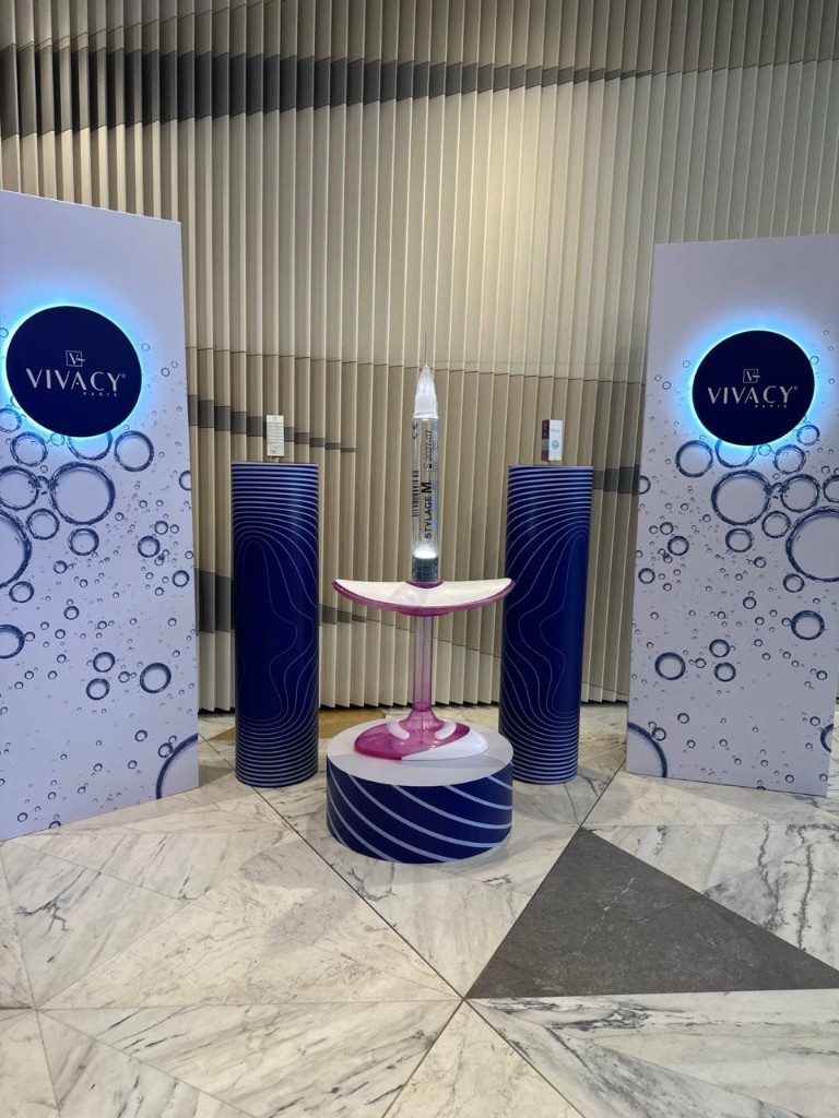 Выставочная фотозона VIVACY PARIS: боковые стенды с подсветкой логотипа, объёмный макет шприца STYLAGE M на подиуме и декоративные колонны